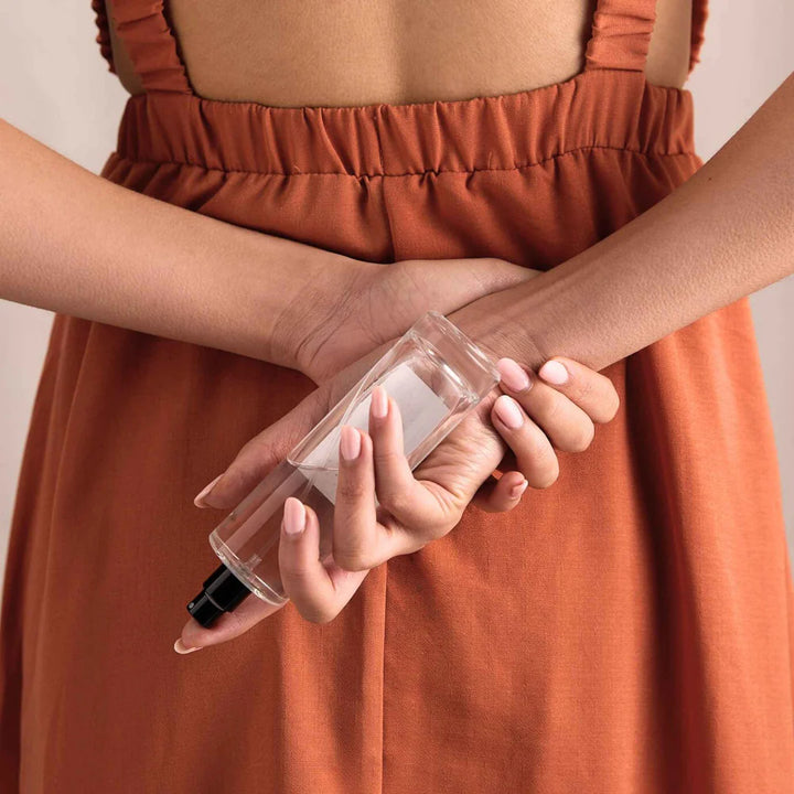 A person wearing a burnt orange dress holding a clear bottle of room spray behind their back