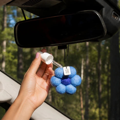 A hand holds a small white dropper bottle above a blue flower-shaped car air freshener with a purple center, hanging in front of a car windshield and rearview mirror. Trees are visible in the background.