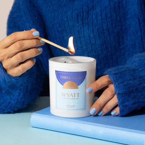 A person in a blue knit sweater holds a lit match above a white candle jar labeled &