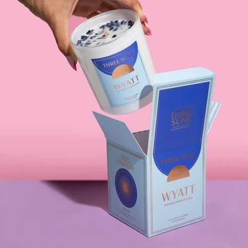 A hand holds a white candle jar above its matching light blue and royal blue box on a pink and lavender studio background. The candle label reads Three Suns Wyatt.