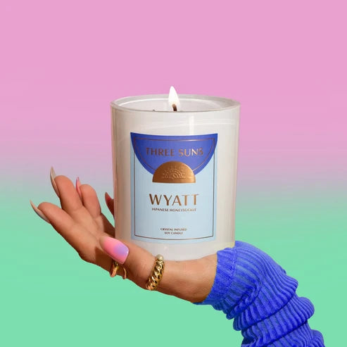 A hand wearing a blue ribbed sweater holds a lit white candle in a clear glass jar. The candle label reads &