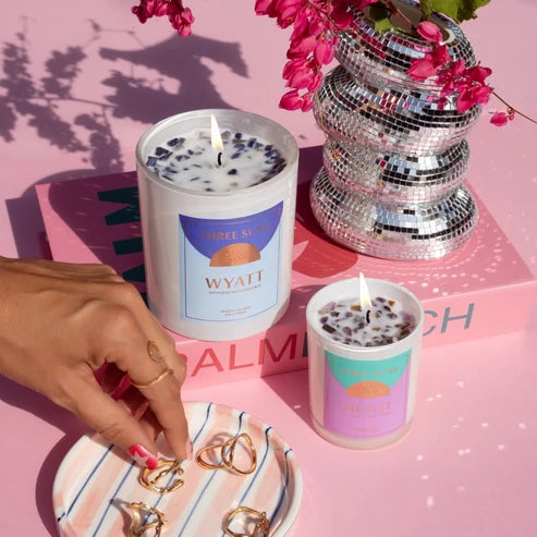 Two lit white soy candles with colorful labels are styled on a pink surface with a pink book, a mirrored disco ball, pink flowers, and a hand placing rings on a striped plate. The larger candle label reads Wyatt and the smaller candle reads Three Suns.