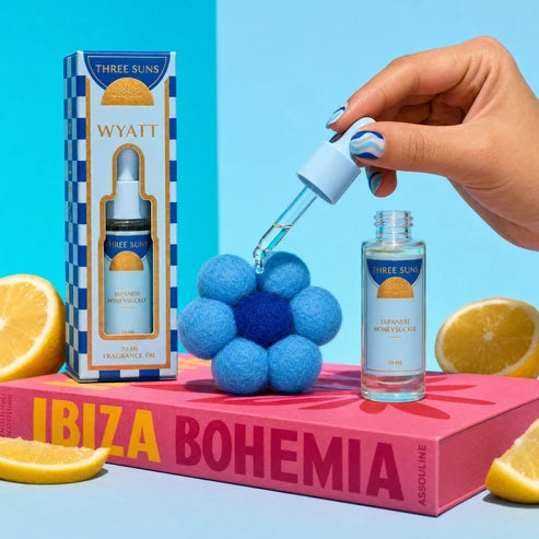 A hand with blue nail art holds a dropper above a small bottle of fragrance oil next to its box and a decorative flower prop, arranged on a pink book with lemon slices against a bright blue background.