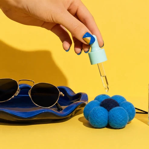 A hand with blue and gold nail polish holds a dropper bottle over a bright yellow surface. The bottle has a pale mint cap and glass pipette, with a clear liquid being dispensed.