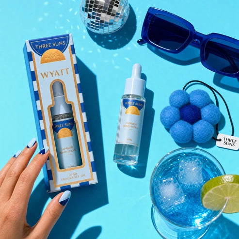 Three Suns Wyatt Fragrance Oil set shown with its retail box, bottle, and dropper on a bright blue styled flat-lay background with sunglasses, a disco ball, a blue flower-shaped hair tie, and a drink with a lime slice. A hand is visible holding the box.