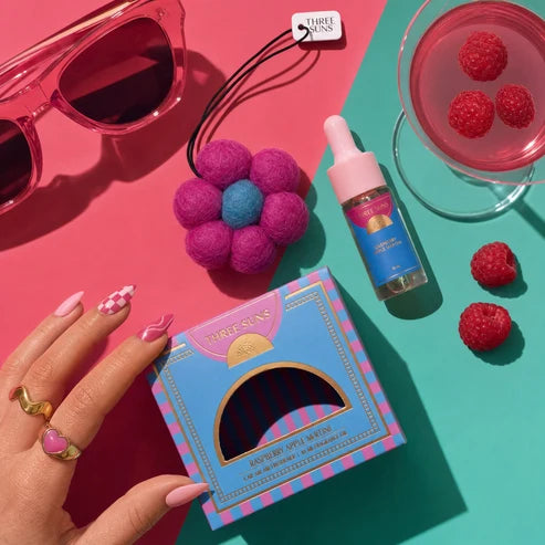 Three Suns Zoi air freshener packaging with a hand holding it, sunglasses, and a drink with raspberries on a colorful background.