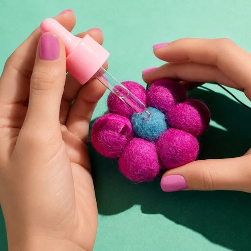 A close-up of hands holding a pink dropper bottle over a small felt flower made of bright pink petals with a blue center, set against a solid teal background.