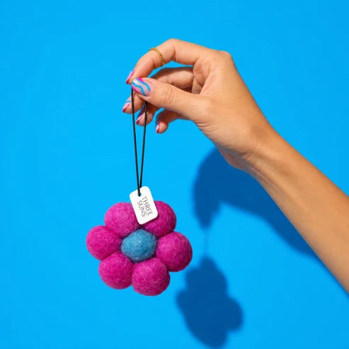 A hand with colorful nail art holds a small felt flower ornament against a bright blue background. The flower has bright pink petals, a blue center, and a small white tag that reads &