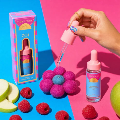 A hand with colorful nail art holds a pink dropper above a small bottle of Three Suns ZOI fragrance oil, with the matching product box standing on a bright blue surface. The scene is styled with raspberries, sliced apple, and a flower-shaped pink object on a pink-and-blue split background.