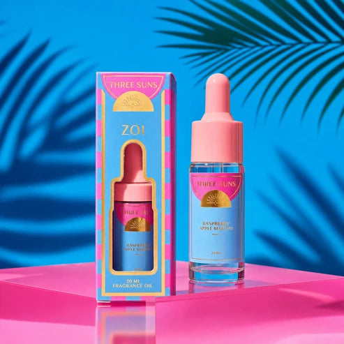 Three Suns Zoi Raspberry Apple fragrance oil bottle and its colorful pink and blue packaging box, set on a vibrant pink reflective surface with a bright blue background and palm leaves.